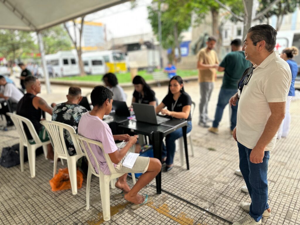 A imagem mostra um ambiente ao ar livre, possivelmente um ponto de registro ou serviço, sob uma grande tenda branca. Várias pessoas estão sentadas em mesas, algumas trabalhando em laptops, enquanto outras estão em pé e observando. Em primeiro plano, um homem com uma camiseta polo clara e jeans azuis está em pé, olhando para a direita, com óculos de sol pendurados na gola da camiseta. À sua esquerda, várias pessoas estão sentadas em mesas pretas. Uma pessoa, vestindo uma camiseta lilás e shorts, está olhando para papéis em suas mãos. Ao lado dela, uma mulher com uma camiseta preta está concentrada em um laptop. Mais ao fundo, outras pessoas estão sentadas e envolvidas com seus laptops. No fundo, há árvores, edifícios e veículos, sugerindo um ambiente urbano ou semiurbano. O chão é pavimentado com um padrão de grade. A atmosfera geral parece ser de atividade organizada.