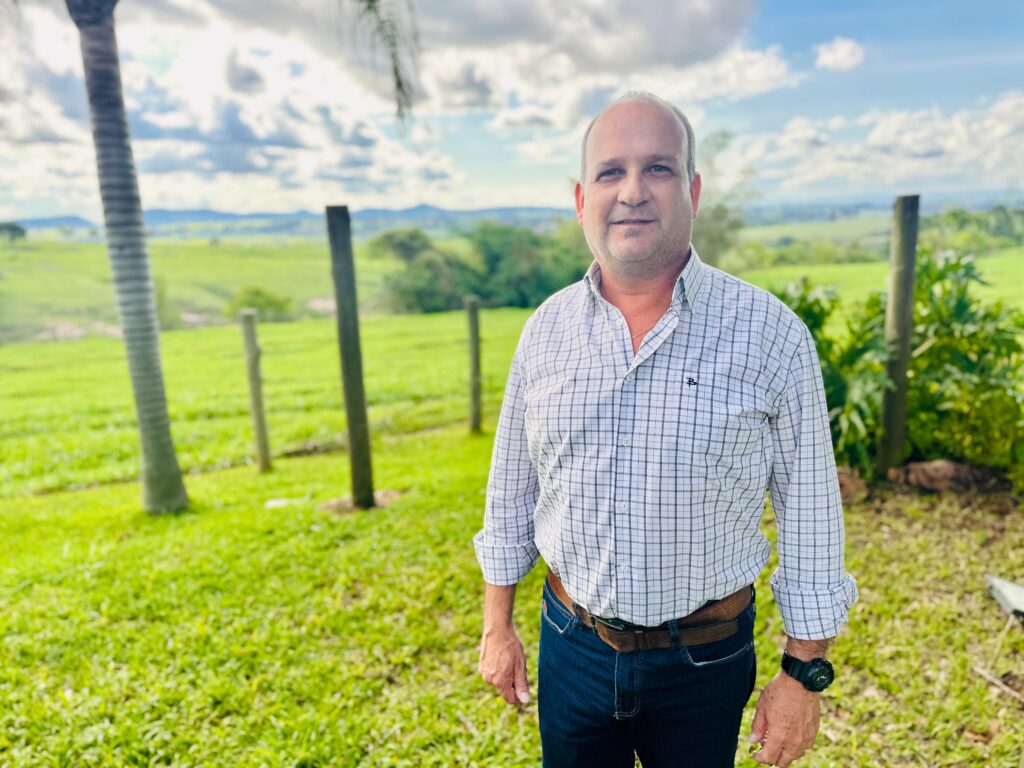 Um homem de meia-idade, com pele clara e cabelo grisalho curto, está em pé em um campo verde. Ele usa uma camisa xadrez de manga comprida, com os punhos dobrados, e calças jeans escuras. Um cinto marrom está preso em sua cintura, e um relógio preto está em seu pulso esquerdo. Ele sorri levemente para a câmera. O fundo é um campo verde exuberante, com algumas árvores e postes de madeira espalhados. Ao longe, colinas suaves se estendem sob um céu azul com nuvens brancas fofas. A luz do sol ilumina a cena, criando uma atmosfera agradável e natural.