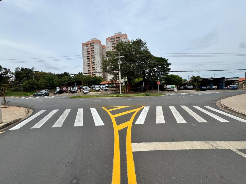 A imagem mostra um cruzamento de ruas com uma faixa de pedestres em primeiro plano. A faixa é composta por barras brancas paralelas pintadas no asfalto, com linhas amarelas em zigue-zague que delimitam a área de travessia. Ao fundo, observam-se prédios residenciais altos com janelas em fileiras. Há também árvores frondosas, postes de luz e fios elétricos cruzando o céu nublado. No lado direito da imagem, há um conjunto de estabelecimentos comerciais, possivelmente oficinas mecânicas ou lojas, com alguns carros estacionados em frente. Um sinal de "Pare" vermelho é visível em um dos cruzamentos. A cena geral sugere um ambiente urbano em um dia com pouca luz solar.