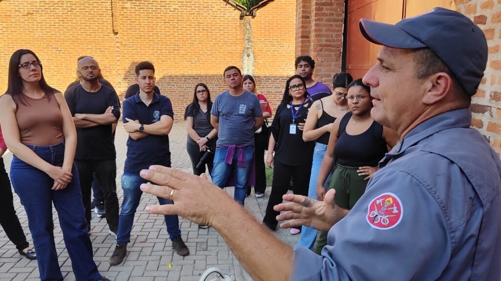 A imagem mostra um grupo de pessoas reunidas do lado de fora, em frente a uma parede de tijolos. Um homem de boné e uniforme cinza, que parece ser um guia ou instrutor, está falando com o grupo e gesticulando com as mãos. Ele tem um distintivo circular com símbolos vermelhos e brancos no ombro de sua camisa. O grupo é composto por homens e mulheres, a maioria olhando atentamente para o homem que fala. Alguns estão de braços cruzados, outros com as mãos juntas. O ambiente parece ser um local público, possivelmente uma instituição ou um ponto turístico com uma arquitetura de tijolos aparente. O chão é pavimentado com pedras em um padrão de paralelepípedos.