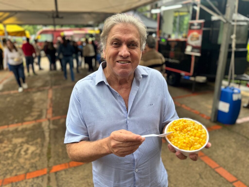 Um homem de cabelos grisalhos e pele clara, vestindo uma camisa azul clara, está sorrindo para a câmera enquanto segura uma tigela branca cheia de milho amarelo com uma colher branca. Ele está em um ambiente externo, com pessoas desfocadas e food trucks ao fundo. O chão é de concreto com marcações de tijolos laranja.