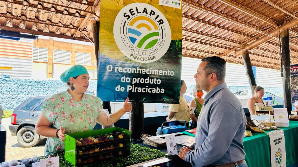 A imagem mostra um evento de venda de produtos locais, com destaque para o selo "SELAPIR - Selo Local de Alimentos Piracicaba". Uma mulher vestida com um avental e um lenço na cabeça, que parece ser uma vendedora, está apresentando seus produtos, possivelmente cupcakes, para um homem. Atrás deles, um grande banner promove o selo e a iniciativa de valorização dos produtos de Piracicaba. O ambiente parece ser uma feira ao ar livre, com uma cobertura de telhas e estruturas de madeira. Outras pessoas e barracas de produtos também são visíveis ao fundo, indicando um evento movimentado.