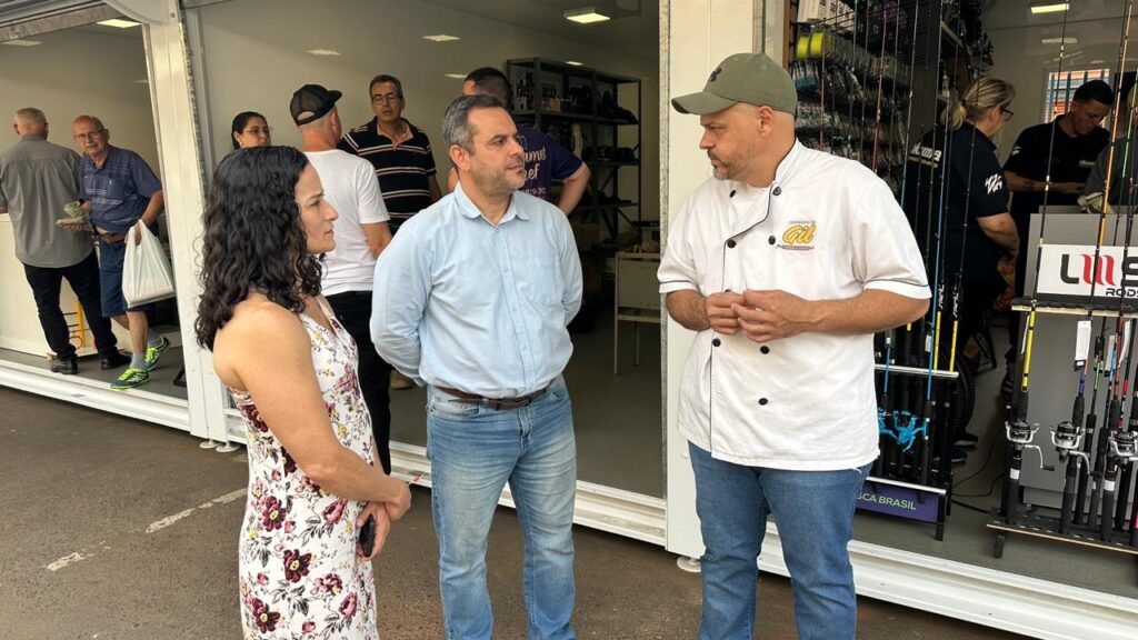 A imagem mostra um grupo de pessoas em frente a uma loja com equipamentos de pesca. No centro, dois homens conversam. Um deles, vestindo uma camisa azul clara e jeans, parece estar explicando algo para o outro, que usa um chapéu verde e um uniforme de chef branco. Atrás deles, outras pessoas observam ou entram na loja. A loja exibe diversas varas de pesca em exposição.