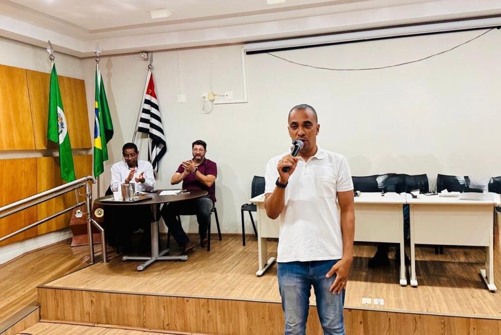O homem em primeiro plano, vestindo uma camisa polo branca e jeans, segura um microfone com a mão direita e fala. Ele está em pé em um palco de madeira, com uma inclinação à esquerda. Atrás dele, sentados em uma mesa redonda, estão dois homens. O homem à esquerda, vestindo uma camisa listrada, aplaude com as mãos. O homem à direita, vestindo uma camisa vinho, também aplaude. Atrás deles, há bandeiras verdes e uma bandeira com listras pretas e brancas. O fundo é uma parede branca com uma tela de projeção e mesas com cadeiras.