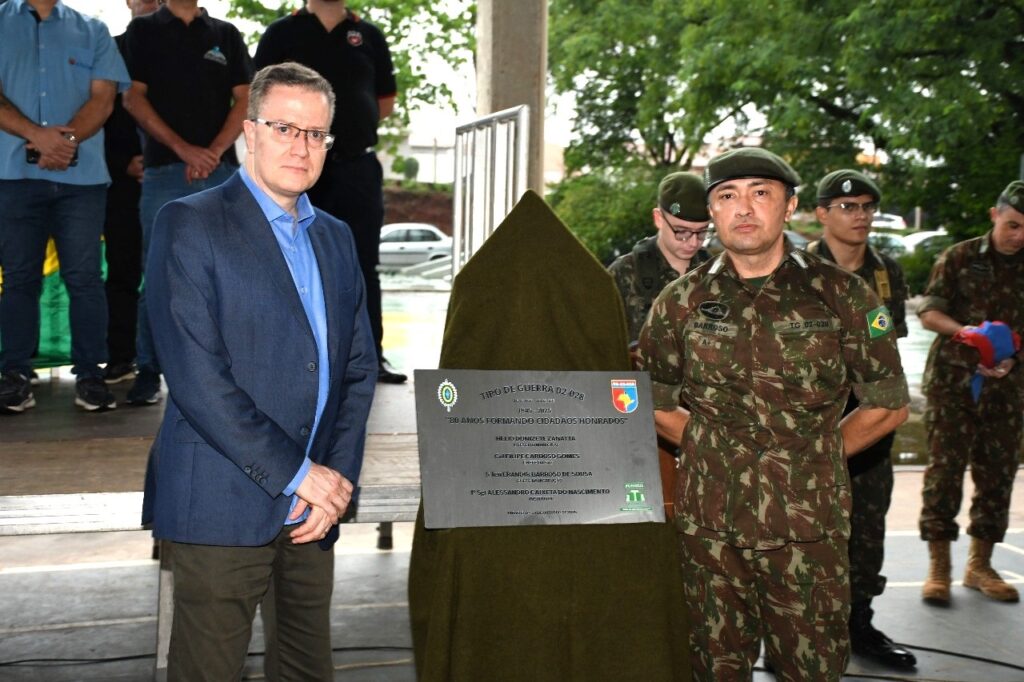 A imagem mostra um evento ao ar livre, possivelmente uma cerimônia militar, com várias pessoas em pé. No centro da imagem, há uma placa cinza com texto em relevo, coberta parcialmente por um tecido verde escuro. A placa comemora os "80 ANOS FORMANDO CIDADÃOS HONRADOS" do "TIRO DE GUERRA 02-028", com as datas de 1945-2025. Abaixo, há nomes e títulos, incluindo "HELIO DONIZETE ZANATTA", "FILIPE CARDOSO GOMES", "ERANDIR BARROSO DE SOUSA" e "ALESSANDRO CAIXETA DO NASCIMENTO". À esquerda da placa, um homem de terno azul escuro e camisa azul clara, com óculos, está de pé e olha para a câmera. Ele tem as mãos cruzadas na frente do corpo. À direita da placa, um homem em uniforme militar camuflado, com um chapéu verde, também está em pé e olha para a frente. Ele tem distintivos no uniforme, incluindo um com a bandeira do Brasil e outro com "TG 02-028". Outros militares em uniformes semelhantes estão atrás dele. Ao fundo, algumas pessoas vestidas com roupas civis e militares podem ser vistas, e há uma estrutura de metal e árvores.