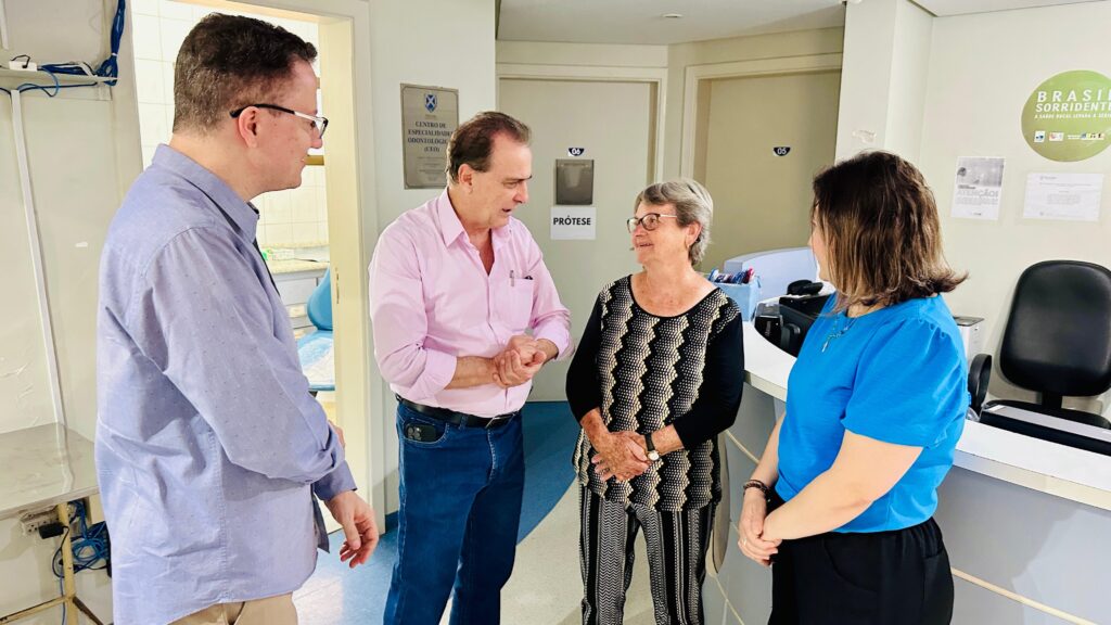 A imagem mostra quatro pessoas em pé em um consultório odontológico, conversando. Duas delas, um homem e uma mulher, estão no centro da imagem, de frente um para o outro. O homem, vestindo uma camisa rosa e jeans, parece estar explicando algo para a mulher, que usa uma blusa preta com um padrão geométrico e calças listradas. À esquerda, um homem com óculos e camisa azul clara está de costas para a câmera, olhando para a cena. À direita, uma mulher com uma blusa azul brilhante e calça preta está de perfil, também observando a conversa. Ao fundo, há portas, um balcão de recepção com materiais de escritório e um cartaz com o logo "BRASIL SORRIDENTE". Há também uma placa com o texto "CENTRO DE ESPECIALIDADE ODONTOLÓGICA (CEO)", indicando que o local é uma clínica especializada em odontologia. As pessoas parecem engajadas em uma conversa profissional ou informativa.