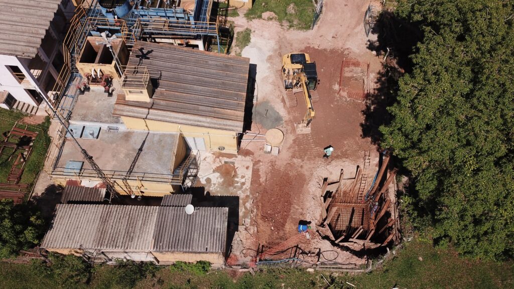 A imagem mostra uma vista aérea de uma área de construção, possivelmente uma estação de tratamento de água ou um local industrial. Vários edifícios e estruturas estão visíveis, incluindo um com telhado de zinco e outro com telhado plano. Uma escavadeira amarela está em operação em uma área de terra batida, onde uma vala está sendo escavada. Há também dois trabalhadores visíveis na área de construção, um perto da escavadeira e outro perto da vala. Uma grande árvore frondosa sombreia parte da cena. A construção parece estar em andamento, com materiais de construção e equipamentos espalhados pela área.