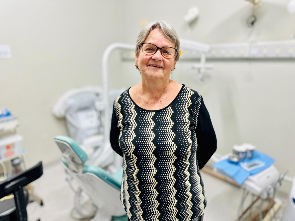 A mulher, com cabelos grisalhos curtos e óculos de aro escuro, sorri para a câmera. Ela está usando uma blusa preta com um padrão em zigue-zague de pontos amarelos e brancos. Seus braços estão cruzados atrás das costas. O fundo sugere que ela está em um consultório odontológico, com uma cadeira odontológica verde e branca visível à sua esquerda e outros equipamentos médicos desfocados ao fundo.