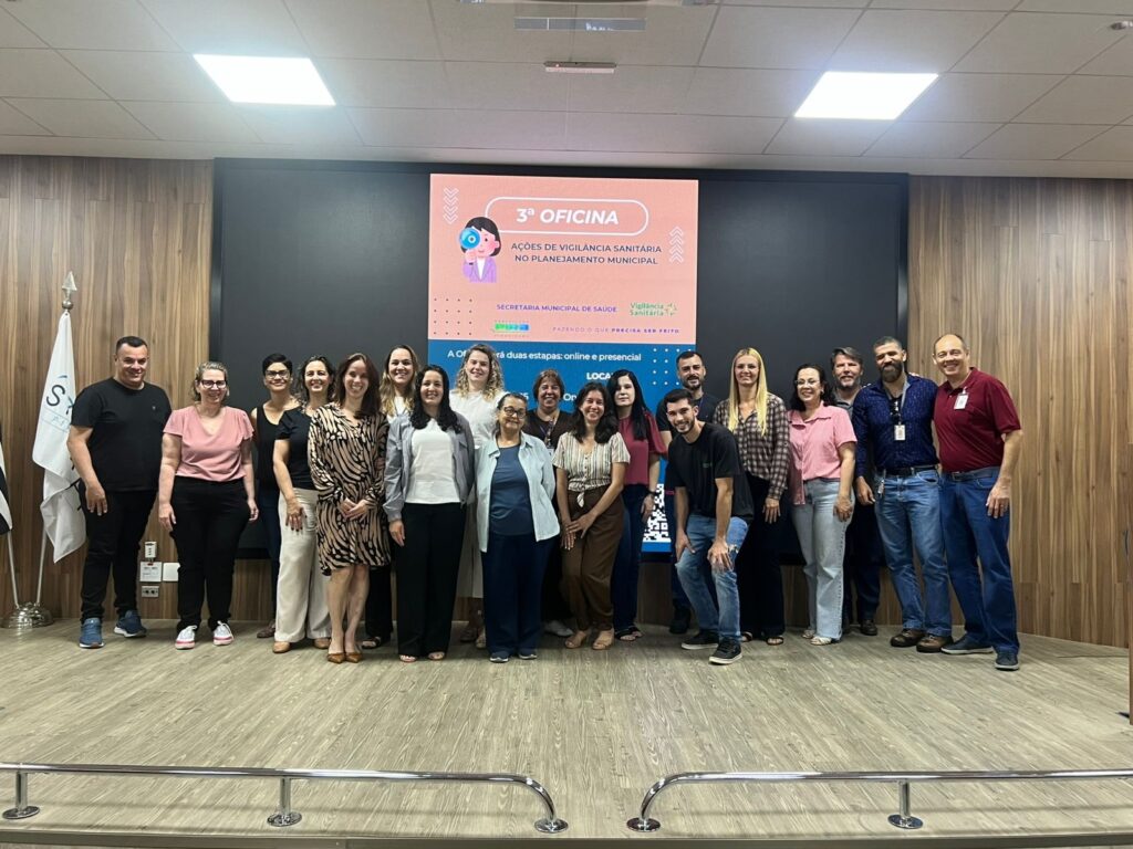A imagem mostra um grupo de pessoas posando para uma foto em frente a um telão. O telão exibe a mensagem "3ª OFICINA AÇÕES DE VIGILÂNCIA SANITÁRIA NO PLANEJAMENTO MUNICIPAL". Abaixo, há logotipos e o texto "SECRETARIA MUNICIPAL DE SAÚDE" e "Vigilância Sanitária". Mais abaixo, lê-se "A Oficina terá duas etapas: online e presencial". As pessoas estão em duas fileiras, algumas em pé e outras agachadas na frente. A maioria veste roupas casuais, com algumas pessoas de camisa social e outras de camiseta. O ambiente parece ser uma sala de conferências ou auditório, com piso de madeira e paredes de madeira escura. Há uma bandeira branca à esquerda da imagem com um logotipo preto e branco.