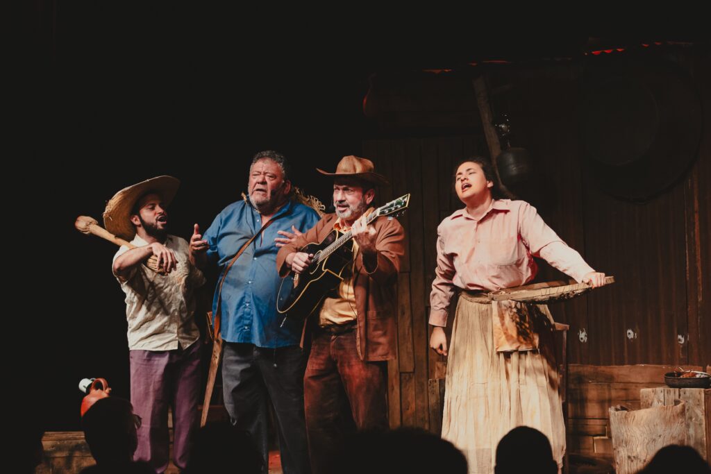 A imagem retrata uma cena teatral com quatro artistas em um palco escuro. No centro, um homem com chapéu de cowboy e jaqueta marrom toca violão, enquanto um homem mais velho, com camisa azul e chapéu de palha, canta expressivamente ao seu lado. À esquerda, um homem com chapéu de palha e camisa clara segura um objeto semelhante a um cajado. À direita, uma mulher com camisa rosa e saia longa também canta com os olhos fechados. O cenário ao fundo sugere um ambiente rústico, com paredes de madeira e elementos suspensos. A iluminação foca nos artistas, criando um contraste dramático com o fundo escuro.