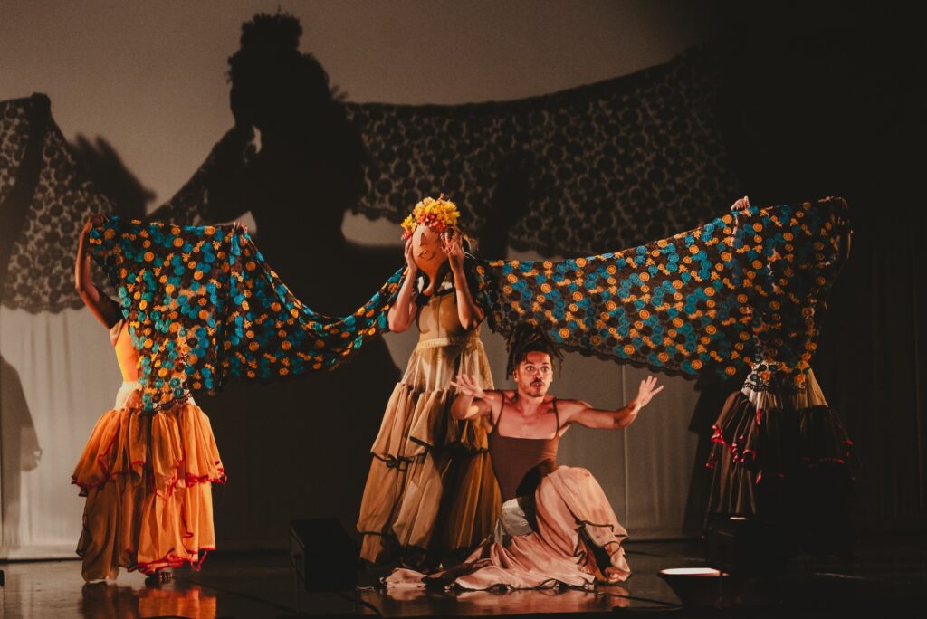 A imagem retrata uma cena de um espetáculo de dança ou teatro, com vários artistas em um palco escuro. No centro, um homem ajoelhado gesticula com as mãos abertas, vestindo uma roupa que parece um maiô marrom e uma saia longa e esvoaçante em tons de rosa e marrom. Atrás dele, uma mulher está em pé, segurando uma máscara em forma de cabaça sobre o rosto, adornada com flores amarelas e laranjas. Ela usa um vestido longo e esvoaçante em tons de bege. Outras duas mulheres, em cada lado da cena, seguram um grande tecido estampado com círculos azuis e laranjas, que se estende horizontalmente, criando um efeito de túnel ou asa. Elas usam vestidos longos e esvoaçantes em tons de laranja e marrom, respectivamente. Ao fundo, uma projeção de uma silhueta feminina com asas se destaca na parede, adicionando profundidade e um toque etéreo à apresentação. A iluminação é focada nos artistas, com o restante do palco em penumbra, realçando a dramaticidade da cena.