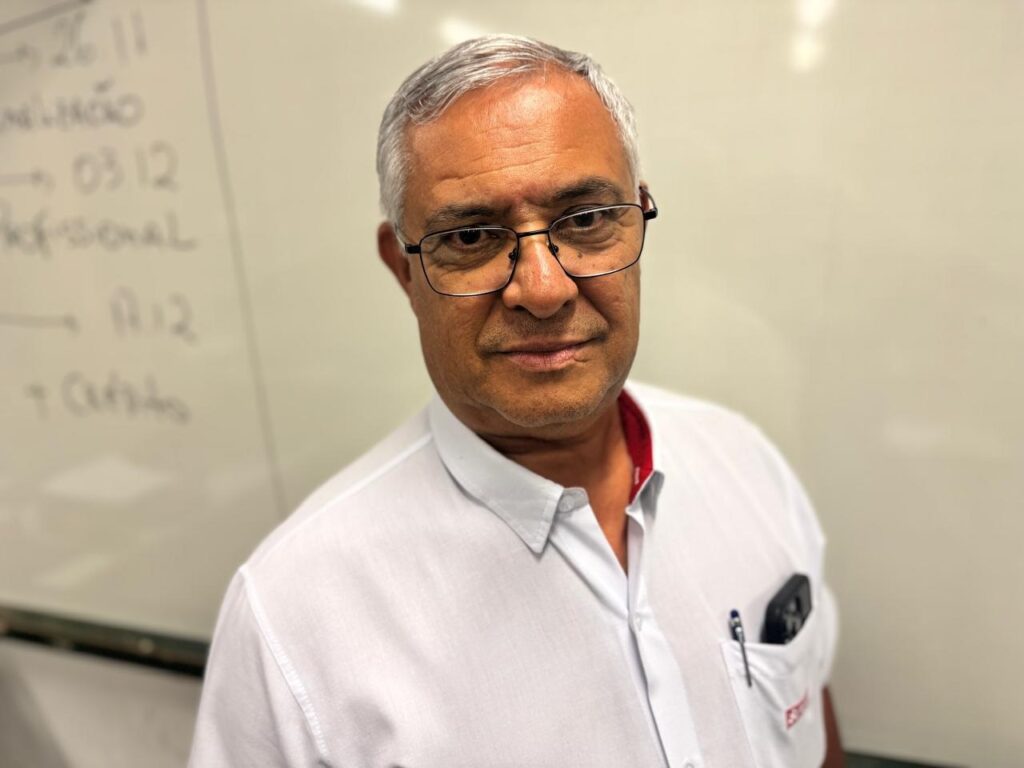 A imagem mostra um homem de meia-idade, com cabelos grisalhos e óculos, olhando diretamente para a câmera. Ele veste uma camisa branca com uma faixa vermelha na gola e tem uma caneta e um objeto preto no bolso da camisa. Ao fundo, há um quadro branco com algumas anotações escritas à mão, incluindo palavras e números como "26 11", "milição", "→ 0312", "Profissional", "A12" e "+ Cursos". O ambiente parece ser um escritório ou uma sala de aula, com iluminação clara e artificial. A expressão do homem é neutra, e a composição da foto foca principalmente no rosto dele, com o quadro ao fundo ligeiramente desfocado.