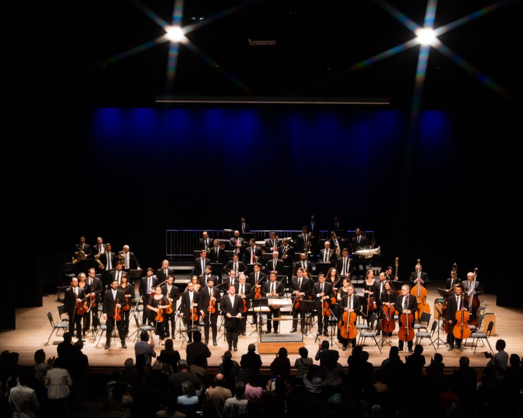 A imagem mostra uma orquestra sinfônica em um palco, com a maioria dos músicos vestindo trajes pretos formais. A orquestra está disposta em fileiras semicirculares, com os instrumentos de corda na frente e os instrumentos de sopro e percussão atrás. O maestro está em pé no centro, de frente para a orquestra. O palco está iluminado por holofotes no alto, que criam um brilho intenso e feixes de luz que se espalham. Ao fundo, há uma cortina azul escura. Na frente do palco, uma plateia de pessoas sentadas em cadeiras assiste à apresentação. A imagem é capturada de um ângulo ligeiramente elevado, oferecendo uma visão completa do palco e da audiência.