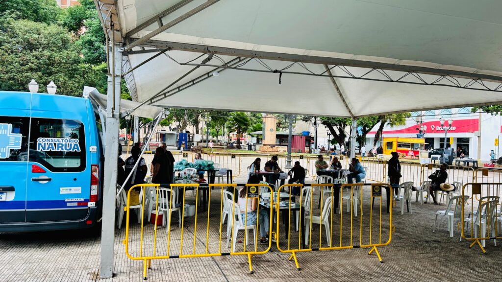 A imagem mostra uma cena ao ar livre, sob uma grande tenda branca, onde várias pessoas estão sentadas à mesa, aparentemente aguardando atendimento ou participando de alguma atividade. Há uma van azul com a inscrição "CONSULTÓRIO N'ARUA" estacionada à esquerda, sugerindo que o local seja um ponto de atendimento móvel. A área sob a tenda está organizada com mesas e cadeiras, e algumas pessoas parecem estar conversando ou preenchendo formulários. A frente da tenda é delimitada por grades amarelas de bloqueio. Ao fundo, observa-se uma praça com árvores, edifícios e outras pessoas circulando, indicando um ambiente urbano movimentado. Em um dos edifícios ao fundo, destaca-se uma fachada vermelha com a inscrição "Bradesco".