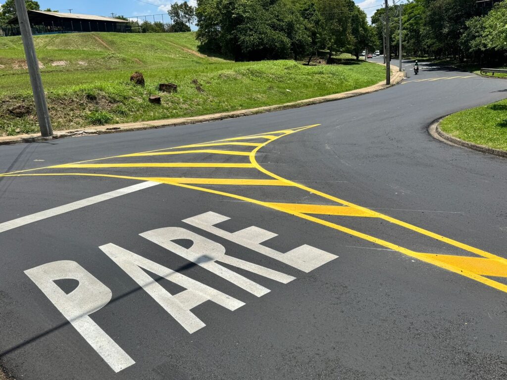 A imagem mostra uma rua asfaltada com sinalização pintada em branco e amarelo. Na parte inferior, em letras brancas grandes, está escrito "PARE", indicando uma parada obrigatória. Ao redor da palavra "PARE", há linhas amarelas em zigue-zague, que parecem delimitar uma área de segurança ou um cruzamento. Essas linhas se estendem pela pista, com uma curva suave à direita. Ao fundo, a rua continua, com uma curva mais acentuada, e é ladeada por grama verde e árvores frondosas. Há também um poste de iluminação pública à esquerda e, mais adiante, um poste de energia elétrica. Em um plano mais distante, veem-se alguns carros e uma motocicleta. O céu está azul com algumas nuvens.