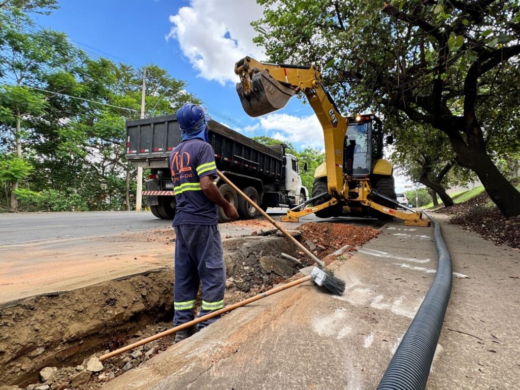 A imagem mostra uma cena de construção ou reparo de infraestrutura em uma área externa, possivelmente urbana ou suburbana. No centro da imagem, um trabalhador vestindo capacete azul, máscara e uniforme de trabalho azul com detalhes em amarelo neon, está em pé ao lado de uma vala aberta. Ele segura um cabo de vassoura e um pedaço de madeira, parecendo estar supervisionando ou auxiliando no trabalho. O uniforme do trabalhador tem um logotipo circular com as letras "RD" e um número de telefone. A vala aberta no chão revela terra e alguns canos de cor cobre ou marrom. Ao fundo, um caminhão basculante preto está estacionado próximo à vala, com a caçamba levantada, sugerindo que material foi ou será despejado. À direita da vala, uma retroescavadeira amarela da marca CAT está posicionada, com seu braço mecânico e caçamba levantados e cheios de terra, indicando que está em operação. As pernas estabilizadoras da máquina estão abaixadas no chão para garantir estabilidade. Um tubo preto corrugado e flexível é visível sobre o concreto na lateral da estrada, estendendo-se para fora da imagem. O cenário geral é de um dia claro, com céu azul pontuado por nuvens brancas e folhagem verde de árvores ao redor, sugerindo um ambiente arborizado. A estrada em si parece ser de asfalto, com a terra da vala contrastando com a superfície.