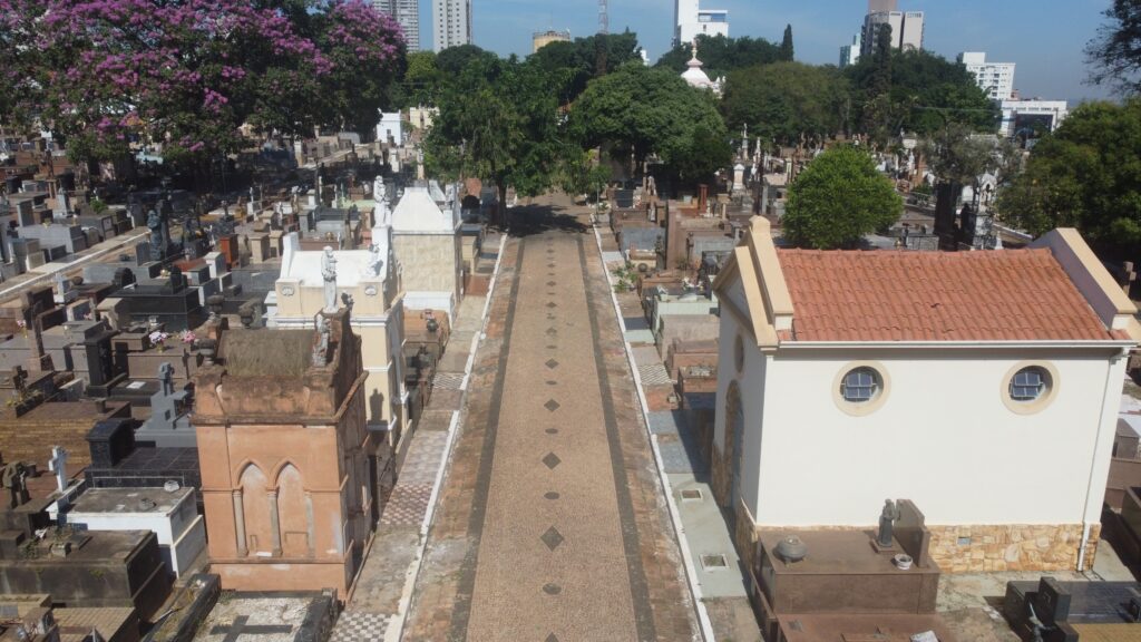 A imagem retrata um cemitério urbano bem cuidado, com uma alameda central pavimentada por ladrilhos decorativos que guia o olhar entre mausoléus e lápides de estilos variados. As construções funerárias exibem uma diversidade arquitetônica — algumas são ornamentadas e imponentes, enquanto outras mantêm linhas simples e discretas. Árvores e vegetação ao fundo suavizam o ambiente, criando um contraste entre o silêncio reverente do espaço e os edifícios modernos que se erguem ao longe, sugerindo a coexistência entre memória e progresso no coração da cidade.