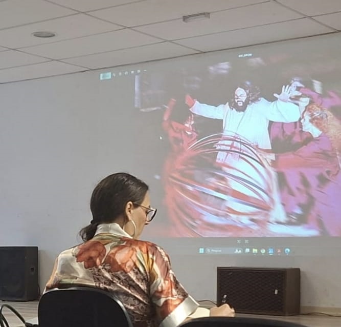 A imagem mostra uma mulher sentada em frente a uma tela de projeção, onde um vídeo está sendo exibido. A mulher está de costas para a câmera, usando um roupão estampado com flores em tons de laranja e bege. Ela tem óculos e seu cabelo está preso em um rabo de cavalo. Na tela de projeção, um homem com barba e cabelo compridos, vestindo uma túnica branca, está no centro, com os braços abertos. Ao redor dele, outras pessoas em trajes escuros parecem estar dançando ou realizando alguma performance, com um grande objeto circular em movimento no centro. A projeção é nítida, mas a imagem da mulher em primeiro plano está um pouco desfocada. Ao lado da mulher, há uma caixa de som preta.