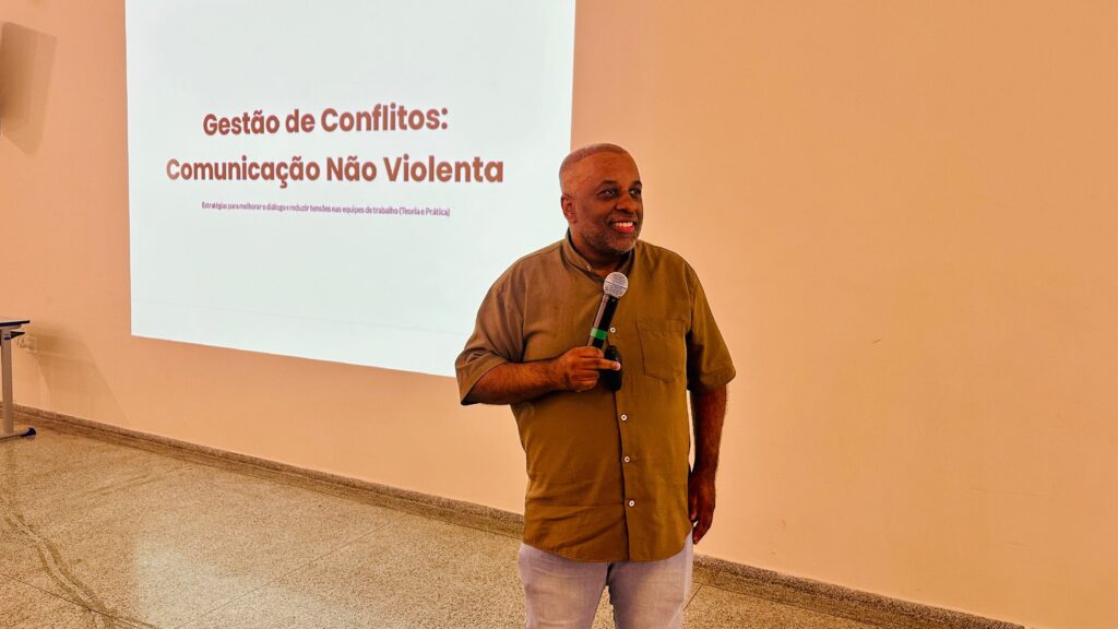 A imagem mostra um homem sorridente, de pele escura, segurando um microfone e falando. Ele está em pé em frente a uma tela de projeção que exibe o título "Gestão de Conflitos: Comunicação Não Violenta" em letras grandes e vermelhas, com um subtítulo em letras menores. O homem veste uma camisa de linho de cor cáqui e calças jeans claras. O fundo da sala é uma parede de cor pêssego e o chão é um piso de granilite. A iluminação é quente, criando uma atmosfera acolhedora.