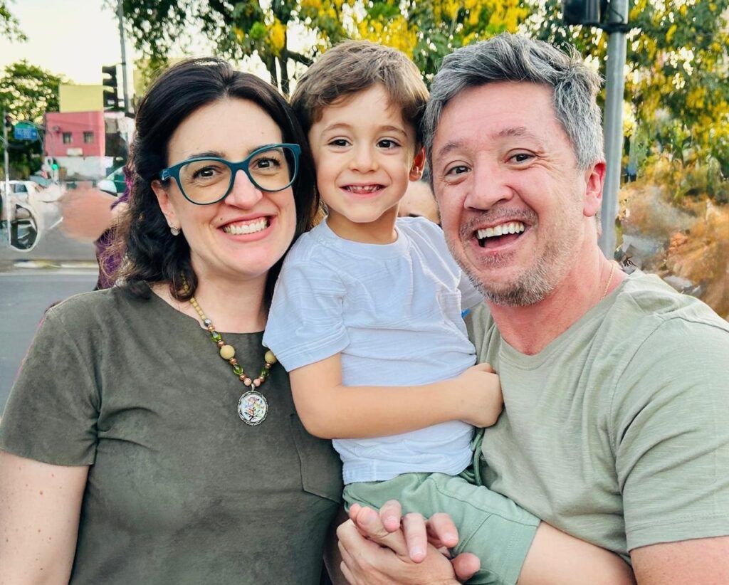 A imagem mostra um casal sorrindo com seu filho no colo, em um momento de alegria e união. A mulher, à esquerda, usa óculos verdes e um colar com pingente redondo. O homem, à direita, tem cabelos grisalhos e segura o menino no colo. O menino, no centro, veste uma camiseta branca e calças verdes, com um sorriso contagiante. O fundo sugere um ambiente externo, com árvores e construções ao longe, indicando um dia ensolarado. A fotografia transmite afeto e felicidade familiar.