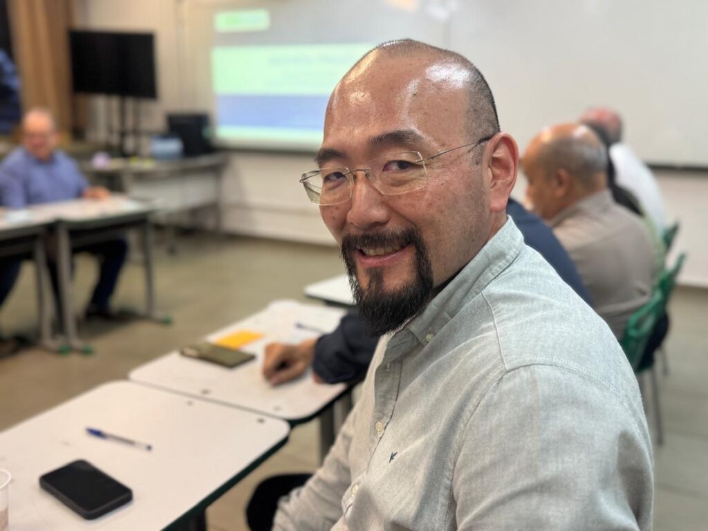 A imagem mostra um homem asiático de meia-idade, careca e com barba e bigode, sorrindo para a câmera. Ele usa óculos e uma camisa cinza clara. Ele está sentado em uma sala de aula, com outras pessoas sentadas em mesas ao fundo. Há um projetor e uma tela ao fundo, sugerindo que uma apresentação está acontecendo. A iluminação é suave e a imagem parece ter sido tirada com um smartphone.