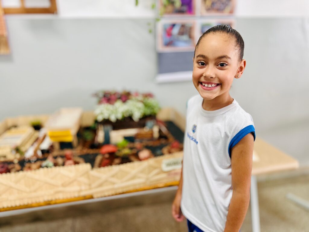 A imagem mostra uma menina sorrindo para a câmera, em primeiro plano. Ela veste uma camiseta branca com detalhes azuis nas mangas e um pequeno emblema no peito. O cabelo dela está preso em um rabo de cavalo. Ao fundo, há uma maquete elaborada, possivelmente um projeto escolar, com elementos que lembram uma paisagem, incluindo plantas em vasos, estruturas que parecem casas e um caminho com cercas de madeira. A iluminação sugere que a foto foi tirada em um ambiente interno, como uma sala de aula.