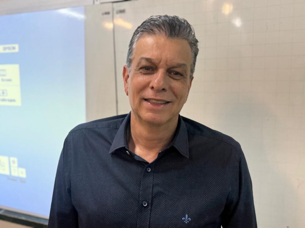 A imagem mostra um homem de meia-idade, com cabelo grisalho penteado para trás, que está sorrindo para a câmera. Ele usa uma camisa escura com um padrão de pequenos pontos brancos e um pequeno bordado de flor de lis no lado esquerdo do peito. O fundo da imagem é dividido. À esquerda, há uma tela de projeção com algumas informações visíveis, incluindo o logotipo da "Epson" e alguns ícones. À direita, há uma parede branca com uma grade quadriculada sutil. A iluminação sugere que a foto foi tirada em um ambiente interno, possivelmente uma sala de aula ou sala de reuniões.