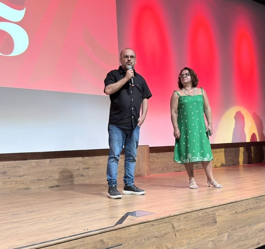 Na imagem, um homem e uma mulher estão em um palco de madeira. O homem, à esquerda, veste uma camisa preta de manga curta e calça jeans azul, segurando um microfone em sua mão direita. Ele tem óculos e está com a mão esquerda no bolso da calça. A mulher, à direita, usa um vestido verde com bolinhas brancas, adornado com um laço em cada alça e uma barra rendada. Ela está descalça, com sandálias brancas nos pés. Ambos olham para a frente, em direção à audiência. O fundo do palco é dominado por uma tela grande com um brilho vermelho e gradiente, com formas arredondadas em tons mais claros de vermelho e amarelo, criando um efeito de iluminação dramático. No canto superior esquerdo da tela, um grande número "5" em branco é visível, possivelmente indicando uma contagem regressiva ou um evento comemorativo. A iluminação geral da cena é suave, destacando os apresentadores no palco.