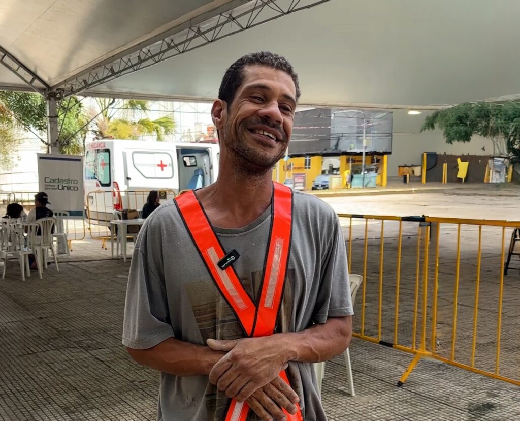 A imagem mostra um homem sorridente, provavelmente de meia-idade, vestindo um colete laranja de alta visibilidade sobre uma camiseta cinza. Ele está com as mãos juntas à frente do corpo e parece estar em um ambiente ao ar livre, sob uma tenda branca. Ao fundo, há uma ambulância branca com uma cruz vermelha e a inscrição "Cadastro Único", indicando que o local pode ser um ponto de atendimento ou evento relacionado a serviços sociais ou de saúde. Também é possível ver algumas cadeiras brancas e pessoas sentadas, além de uma barreira amarela, sugerindo organização do espaço para atendimento ao público. O cenário ao fundo apresenta construções e uma atmosfera urbana. A expressão do homem transmite simpatia e alegria, e o ambiente aparenta ser um local de apoio comunitário, possivelmente no Brasil, considerando as referências presentes.