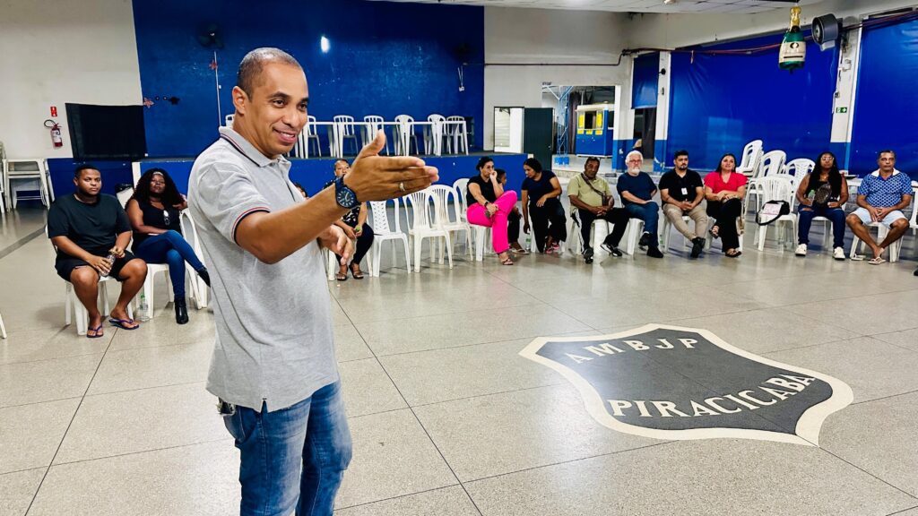 A imagem mostra um grupo de pessoas sentadas em cadeiras brancas dispostas em um círculo, com um homem em primeiro plano gesticulando enquanto fala. O ambiente parece ser um salão com paredes azuis e brancas, e um grande emblema no chão com as inscrições "AMBJP PIRACICABA". Há uma garrafa de espumante pendurada no teto. As pessoas sentadas vestem roupas casuais e parecem estar participando de uma reunião ou palestra.