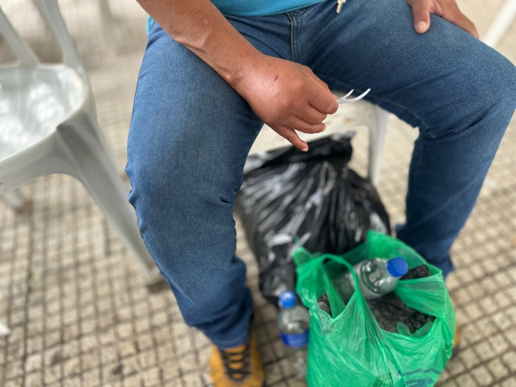 A imagem mostra uma pessoa sentada em uma cadeira de plástico branca, vestindo calça jeans azul e uma camiseta azul. A pessoa está segurando um pequeno objeto branco na mão direita. Ao lado da pessoa, no chão, há um saco de lixo preto e uma sacola verde contendo garrafas de água e outros itens. O chão é coberto por um padrão de azulejos quadrados em tons de bege e marrom. A iluminação sugere que a foto foi tirada durante o dia, possivelmente em um ambiente externo ou em uma área com luz natural.