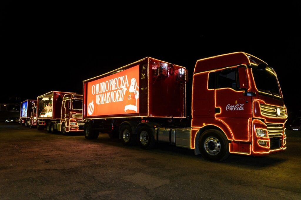 A imagem exibe uma fila de caminhões da Coca-Cola decorados com luzes de Natal, estacionados em um local escuro, possivelmente à noite. O caminhão da frente, em destaque, é vermelho e adornado com fileiras de luzes amarelas que contornam sua cabine e reboque. O reboque possui um grande painel luminoso com a mensagem em português "O MUNDO PRECISA DE MAIS NOEIS", acompanhada de uma ilustração de Papai Noel. Os outros caminhões na fila também são decorados com luzes, embora os detalhes sejam menos nítidos. O fundo é completamente preto, contrastando fortemente com as luzes brilhantes dos caminhões, criando uma atmosfera festiva e mágica. A iluminação dos caminhões realça a identidade visual da Coca-Cola, com seu vermelho característico e o logotipo proeminente.