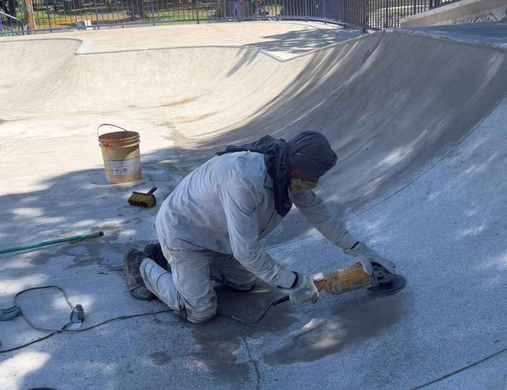 A imagem mostra um trabalhador ajoelhado dentro de uma piscina de skate de concreto, usando uma esmerilhadeira para alisar a superfície. Ele está usando roupas de proteção, incluindo um lenço na cabeça, máscara e luvas, e a área ao redor dele está molhada. Há um balde de tinta velho e uma escova perto dele, e um tubo verde (possivelmente uma mangueira) se estende para a esquerda. A piscina de skate parece estar em construção ou em processo de renovação, com a superfície de concreto visivelmente sendo trabalhada. A luz do sol projeta sombras na área.