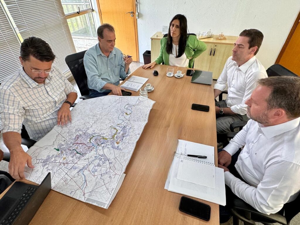 A imagem mostra um grupo de quatro pessoas sentadas ao redor de uma mesa de madeira, aparentemente em uma reunião de trabalho. À esquerda, um homem de camisa xadrez e óculos está debruçado sobre um mapa grande e detalhado, apontando para ele com a mão. Ao lado dele, um homem de camisa azul clara segura uma caneta e olha para um caderno aberto à sua frente. Do outro lado da mesa, uma mulher com uma jaqueta verde e um homem de camisa branca estão sentados, observando algo. Há também um laptop, telefones celulares, xícaras de café e pilhas de papéis sobre a mesa. O ambiente parece ser um escritório, com janelas e armários ao fundo.