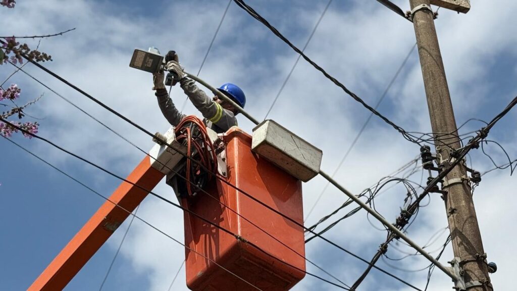 A imagem mostra um trabalhador em uma cesta de elevador laranja, subindo em direção a fios elétricos. Ele está usando um capacete azul e luvas, e parece estar ajustando um aparelho preso a um dos fios. Ao fundo, o céu está parcialmente nublado e uma árvore com flores roxas pode ser vista à esquerda. Um poste de concreto com vários fios e isoladores elétricos também está presente na cena. A perspectiva da foto é de baixo para cima, destacando a altura do trabalhador e a complexidade da rede elétrica.