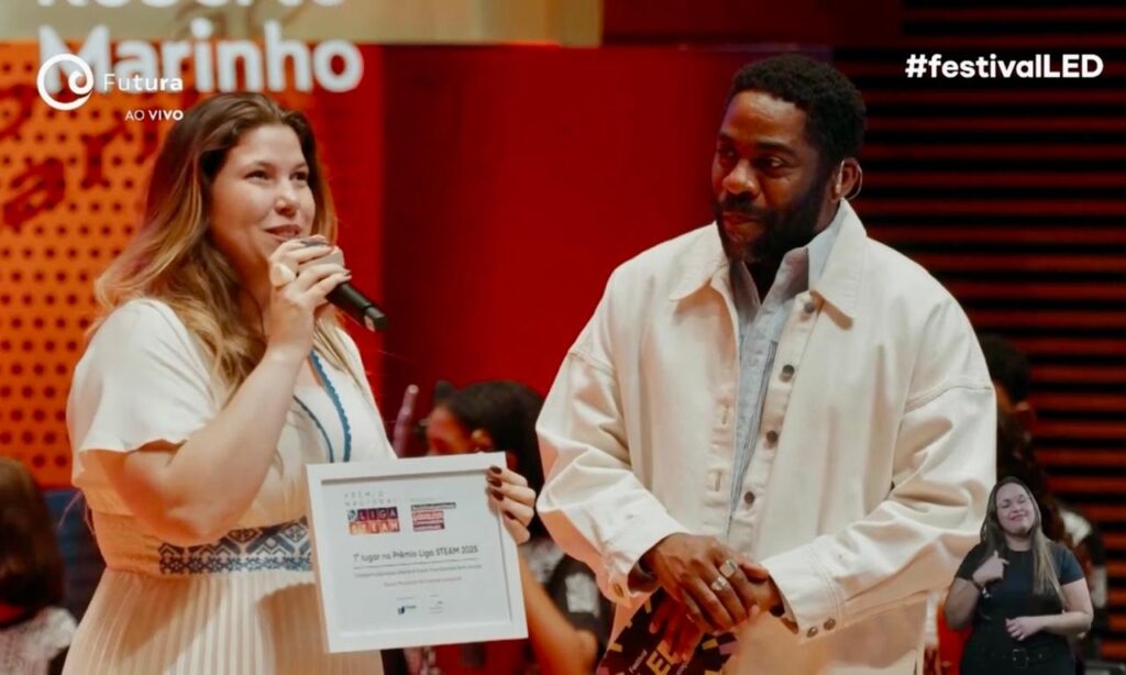 A imagem mostra um homem e uma mulher em um palco. A mulher, à esquerda, está segurando um microfone e um certificado. Ela usa um vestido branco com detalhes azuis na gola e na cintura. O homem, à direita, veste uma camisa clara e um casaco bege. Ele tem barba e usa anéis nas mãos. Ao fundo, há uma parede vermelha com listras horizontais e um letreiro com "Futura AO VIVO" e o logotipo da emissora. No canto superior direito, aparece a hashtag "#festivalLED". Há também uma pequena janela com uma mulher interpretando em Libras.