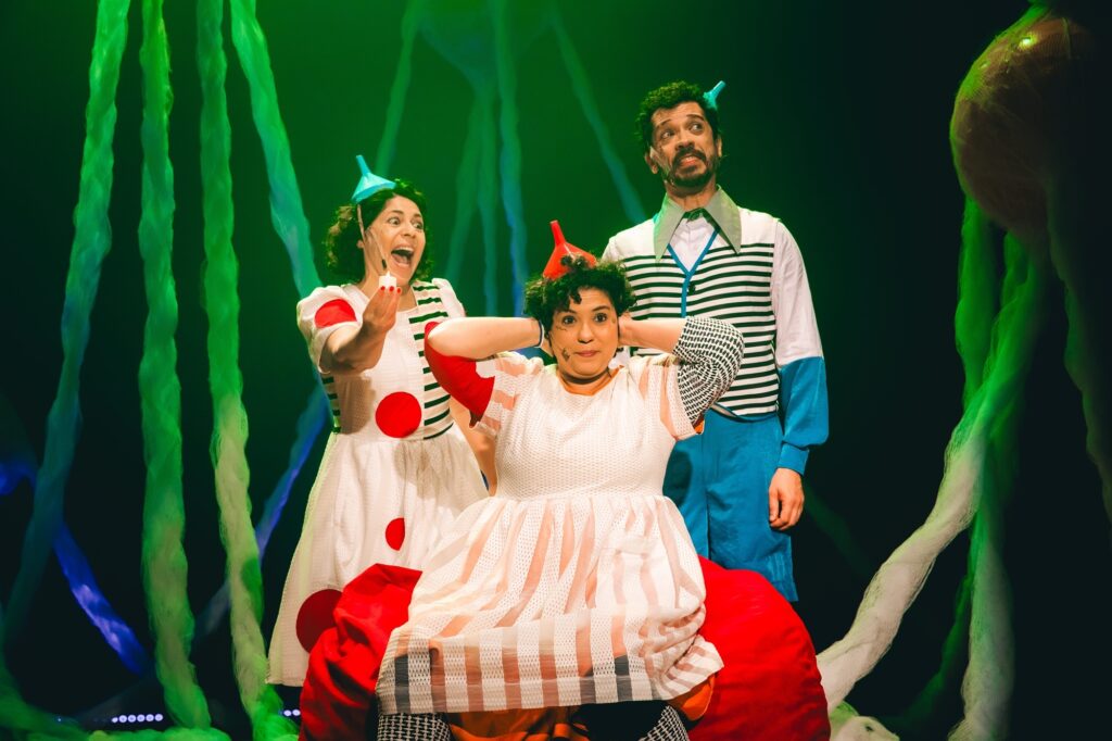 A imagem mostra três atores em um palco, com um cenário que sugere um ambiente subaquático ou alienígena, com longos tentáculos verdes pendurados. No centro, uma atriz com cabelo cacheado escuro, usando um vestido branco com listras cor de pêssego e um chapéu em formato de funil vermelho, cobre os ouvidos com as mãos, com uma expressão de surpresa ou desconforto. À esquerda, outra atriz, com cabelo escuro preso, veste um vestido branco com grandes bolinhas vermelhas e um chapéu funil azul. Ela segura um pequeno objeto branco e tem a boca aberta em uma expressão de espanto ou alegria. À direita, um ator com barba escura e cabelo curto, usa uma roupa vibrante com um colete listrado em preto e branco sobre uma camisa branca, calças azuis e um colarinho verde. Ele olha para cima com uma expressão intrigada. A iluminação é dramática, com tons verdes predominando no fundo e focando nos atores, criando uma atmosfera teatral e lúdica.