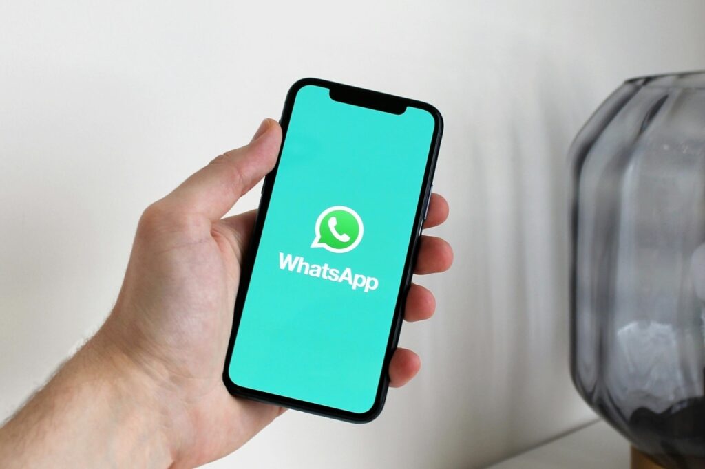A imagem mostra uma mão segurando um smartphone com a tela ligada. A tela exibe o ícone e o nome do aplicativo WhatsApp em um fundo verde-água vibrante. O ícone do WhatsApp é um círculo branco com um telefone dentro, sobreposto a uma bolha de fala branca. O nome "WhatsApp" está escrito em letras brancas abaixo do ícone. O smartphone parece ser um modelo moderno com uma tela de ponta a ponta e uma câmera frontal proeminente na parte superior. Ao lado do telefone, há um vaso de vidro cinza escuro e um fundo branco claro, criando um ambiente limpo e minimalista.