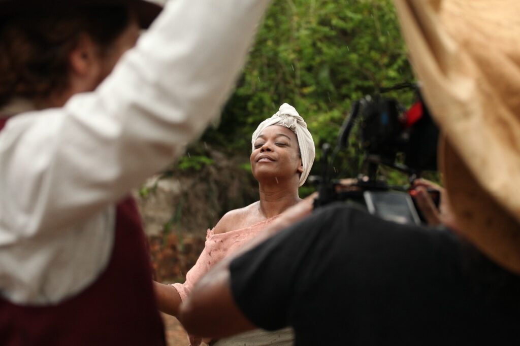 A imagem mostra uma mulher negra em um close médio, com os olhos fechados e uma expressão serena no rosto. Ela usa uma faixa branca na cabeça e uma roupa rosa clara que deixa os ombros à mostra. A cena é ao ar livre, com um fundo desfocado de folhagens verdes e um pouco de parede de tijolos, sugerindo um ambiente natural ou rústico. Está chovendo levemente, com gotas visíveis na pele da mulher e no ar. Ao redor dela, aparecem duas pessoas parcialmente visíveis: à esquerda, alguém com camisa branca, colete escuro e chapéu, com o braço levantado; à direita, outra pessoa usando chapéu e camiseta preta, segurando uma câmera de vídeo, indicando que se trata de uma gravação ou filmagem. A iluminação é suave e difusa, típica de dias nublados ou chuvosos, destacando a pele molhada da mulher. O foco principal está nela, enquanto as outras figuras e o fundo estão desfocados, direcionando a atenção para sua expressão calma e introspectiva. A imagem transmite uma sensação de paz e reflexão em meio à chuva, possivelmente capturada durante uma produção audiovisual.