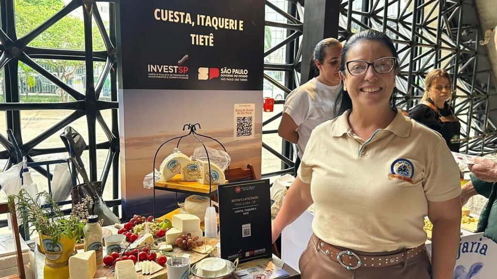 A imagem mostra uma mulher sorrindo para a câmera em um evento, com uma mesa cheia de queijos e acompanhamentos à sua frente. Ela usa uma camisa bege com um emblema no peito e um cinto marrom com uma fivela decorada. Atrás dela, há um banner com o título "CUESTA, ITAQUERI E TIETÊ" e os logotipos da INVESTSP e do Governo do Estado de São Paulo. A mesa exibe uma variedade de queijos, uvas, tomates cereja e outros alimentos, dispostos de forma atraente. Outras pessoas são vistas ao fundo, indicando um evento ou feira. O ambiente parece ser interno, com uma estrutura metálica visível no canto superior esquerdo.