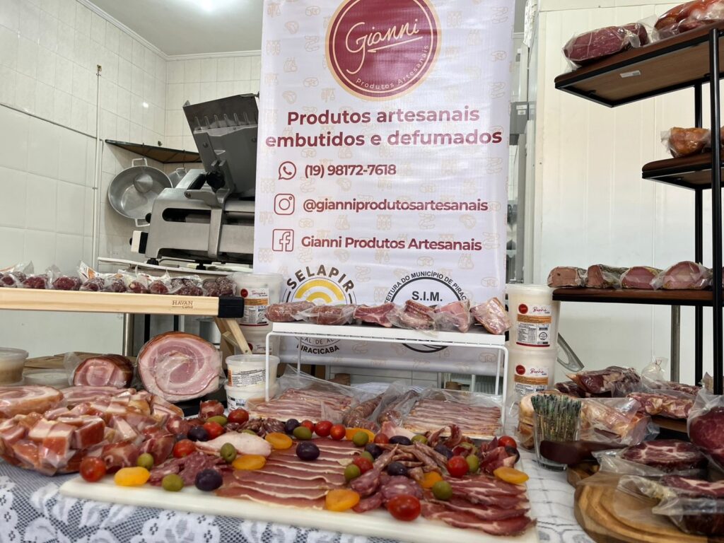 A imagem exibe uma mesa repleta de produtos artesanais de carne, como salames, presuntos, bacon e outras carnes defumadas e embutidas. Há também frutas como uvas e damascos, que parecem acompanhar os produtos de carne em uma tábua de frios. Ao fundo, um banner da marca "Gianni Produtos Artesanais" apresenta o nome da empresa, telefone e redes sociais. Uma máquina fatiadora profissional está visível à esquerda, sugerindo um ambiente de produção ou venda de carnes. Prateleiras com mais produtos de carne e potes de armazenamento completam a cena, que transmite a ideia de um negócio especializado em carnes de qualidade.