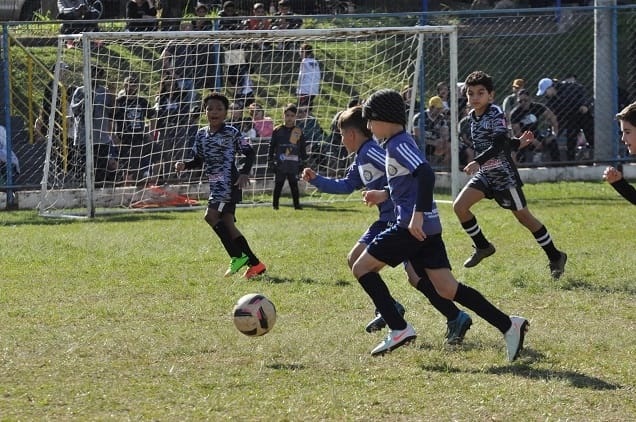 A imagem mostra uma partida de futebol juvenil em andamento, com vários garotos correndo atrás da bola em um campo gramado. O foco está em cinco jogadores que estão em movimento, alguns vestindo uniformes azuis e outros com uniformes pretos e brancos. A bola está próxima ao grupo, ligeiramente à esquerda do centro da imagem. Ao fundo, é possível ver o gol com a rede e algumas pessoas assistindo ao jogo. O ambiente é ao ar livre, com iluminação natural, sugerindo que é durante o dia. A cena transmite energia e ação, típica de um momento competitivo em uma partida de futebol entre crianças.
