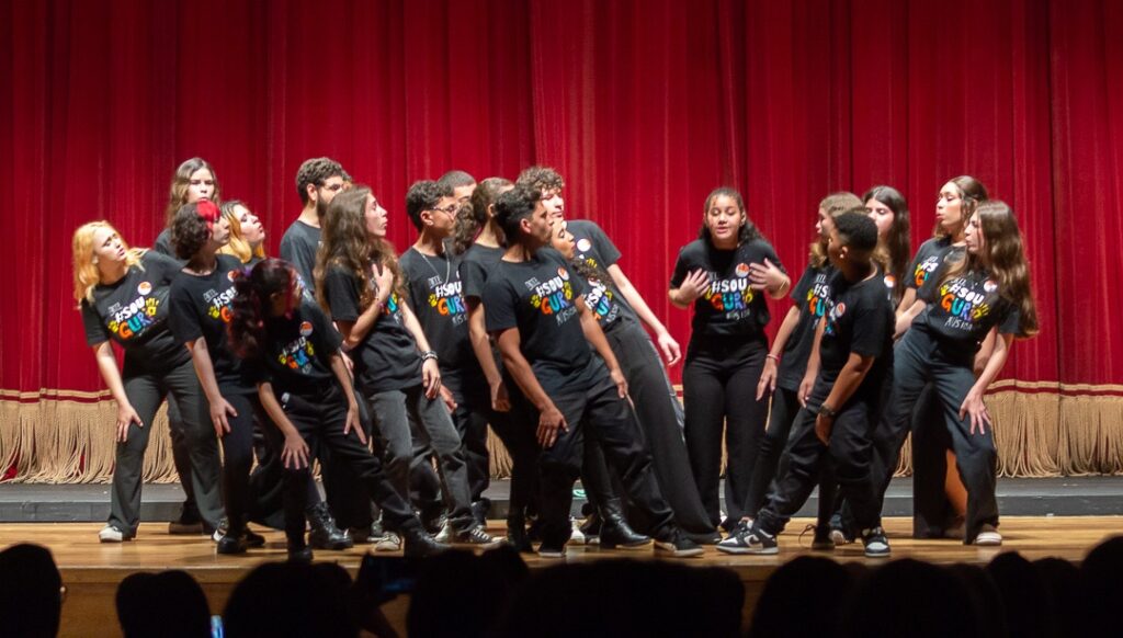 A imagem mostra um grupo de jovens em um palco, vestindo camisetas pretas idênticas com a inscrição "# SOU GURI" estampada em cores vibrantes. Eles estão posicionados em frente a uma cortina vermelha e parecem estar realizando uma performance, possivelmente uma dança ou uma apresentação teatral, dada a pose inclinada e expressiva de muitos deles. As luzes do palco iluminam o grupo, destacando suas ações e vestimentas. Há um público na parte inferior da imagem, cujas cabeças desfocadas indicam que estão assistindo à apresentação. As camisetas com a mensagem "# SOU GURI" sugerem que o grupo faz parte de uma iniciativa ou projeto com esse nome.