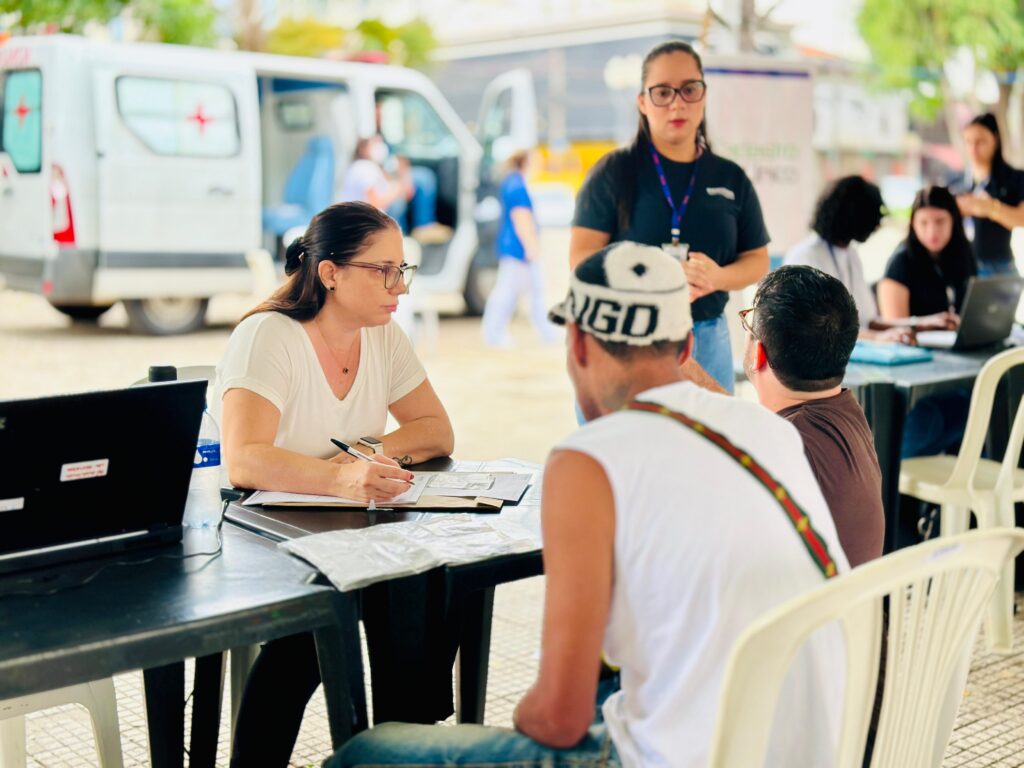 A imagem retrata uma cena ao ar livre, possivelmente um posto de atendimento ou evento de saúde, com várias pessoas interagindo. No primeiro plano, à esquerda, uma mulher com óculos e uma blusa branca está sentada a uma mesa escura, escrevendo em papéis com uma caneta. Ao seu lado, um laptop preto está aberto. À sua frente, um homem com um boné branco e preto com a inscrição "NGO" e uma camiseta branca está de costas, olhando para a mulher. Perto dele, outro homem com uma camiseta marrom também está sentado, de costas para a câmera. Ao fundo, uma van branca com uma cruz vermelha visível na porta traseira aberta sugere um contexto médico ou de emergência. Uma mulher com óculos e uma camiseta preta, usando um crachá, está em pé atrás da mesa, olhando para os lados. Outras pessoas estão visíveis em segundo plano, algumas sentadas em mesas com laptops, e outras em movimento. O ambiente parece ser um espaço aberto, com iluminação natural e um piso quadriculado.