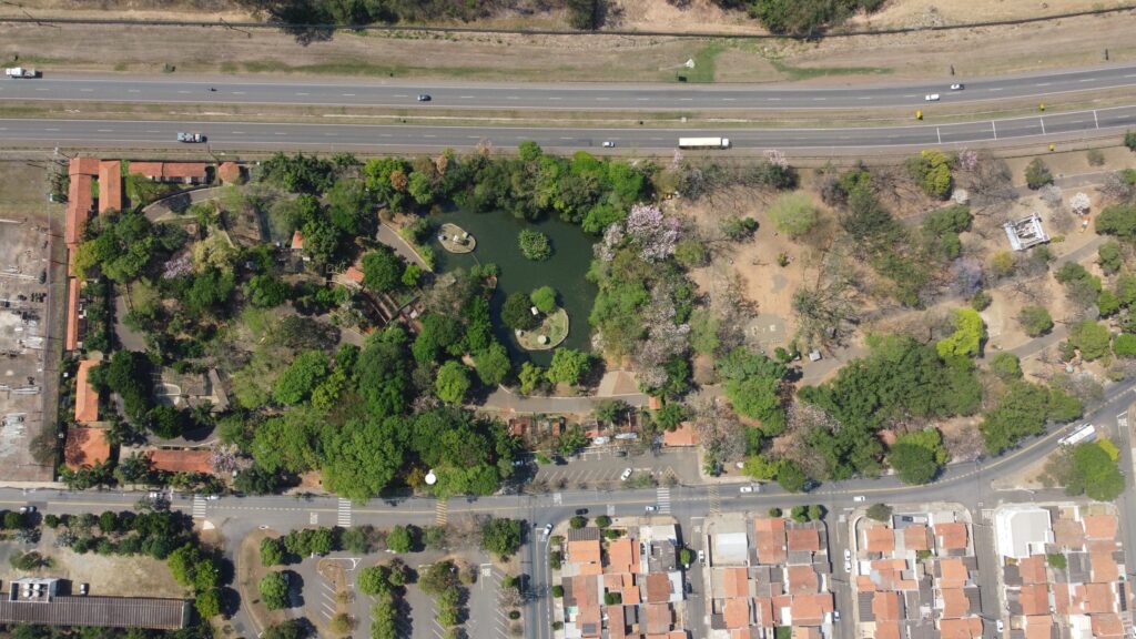 A imagem apresenta uma vista aérea de uma área urbana com destaque para um grande parque arborizado que abriga um lago com pequenas ilhas, onde se observam estruturas que parecem gazebos ou elementos decorativos. O parque é cercado por bairros residenciais com telhados vermelhos, vias movimentadas com veículos e algumas construções comerciais ou institucionais. Caminhos sinuosos e espaços abertos indicam áreas destinadas ao lazer e à circulação de visitantes.