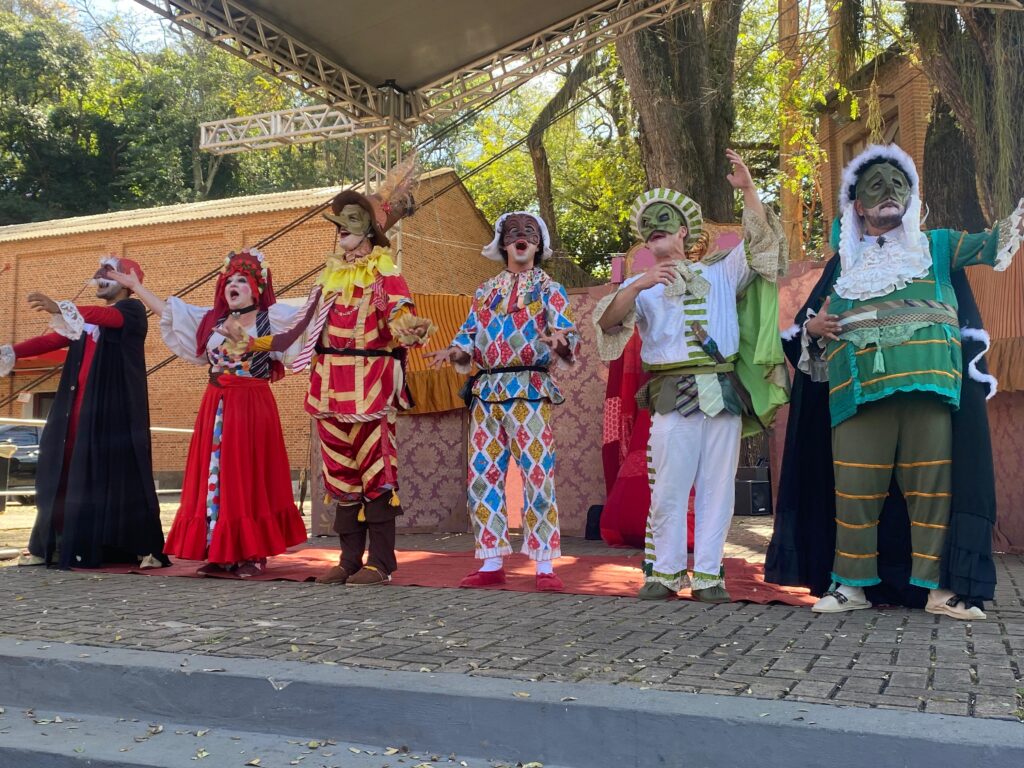A imagem retrata um grupo de seis artistas mascarados em um palco ao ar livre, com uma estrutura metálica de palco acima e árvores e um edifício de tijolos ao fundo. Todos os artistas usam trajes coloridos e elaborados, com máscaras que cobrem seus rostos. Eles parecem estar no meio de uma performance, com poses dramáticas e expressões faciais exageradas através das máscaras. Da esquerda para a direita: * O primeiro artista usa um manto preto sobre um traje vermelho com gola branca rendada. * O segundo artista veste um traje vermelho vibrante com detalhes em dourado e uma saia longa. * O terceiro artista está em um traje vermelho e amarelo em um padrão xadrez, com uma máscara marrom e um adorno de penas na cabeça. * O quarto artista está em um traje vibrante com um padrão de losangos coloridos em azul, vermelho, amarelo e branco, e uma máscara branca. * O quinto artista usa um traje branco com detalhes em verde e várias gravatas coloridas em volta do pescoço e cintura. * O sexto artista está em um traje verde escuro com detalhes em dourado e azul, uma capa preta e uma máscara verde com uma gola branca volumosa. A iluminação sugere que é um dia ensolarado. O palco está montado em uma área pavimentada, com algumas folhas caídas no chão.