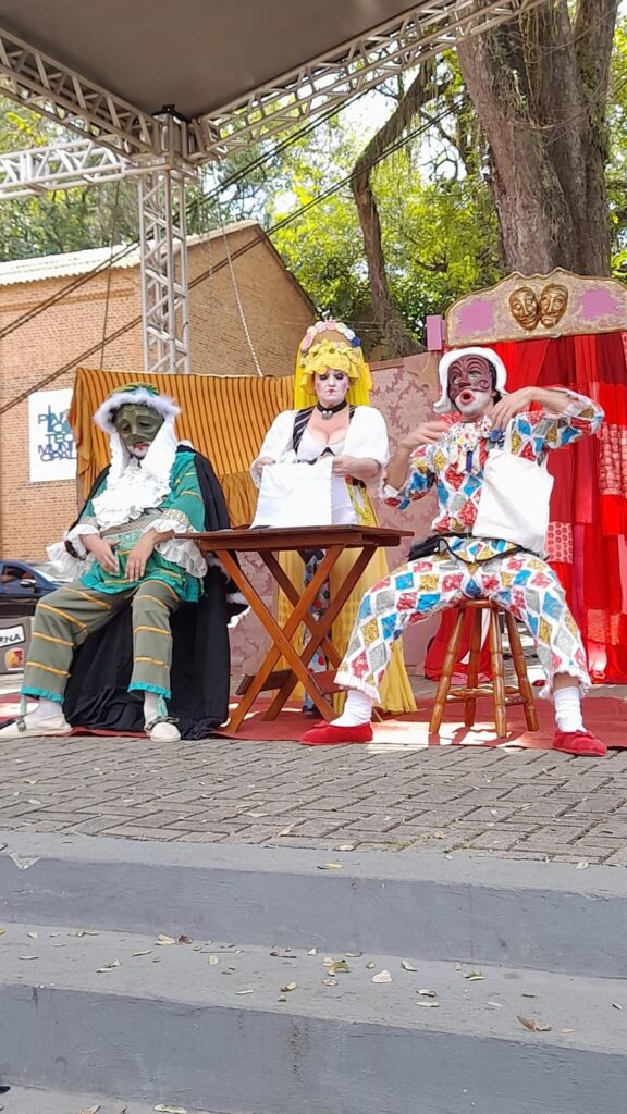 A imagem mostra uma performance teatral ao ar livre, com três atores em figurinos elaborados. No lado esquerdo, um ator com uma máscara verde e um traje verde e branco com detalhes em azul está sentado, vestindo uma capa preta. No centro, uma atriz com um chapéu amarelo volumoso e um vestido branco com decote em V está em pé atrás de uma mesa de madeira, segurando um objeto branco. À direita, um ator com uma máscara branca e vermelha e um traje colorido de losangos está sentado em um banquinho, gesticulando com as mãos. O fundo é decorado com cortinas vermelhas e um painel com máscaras teatrais. O palco parece ser montado em um espaço público, com uma estrutura metálica e árvores ao fundo.