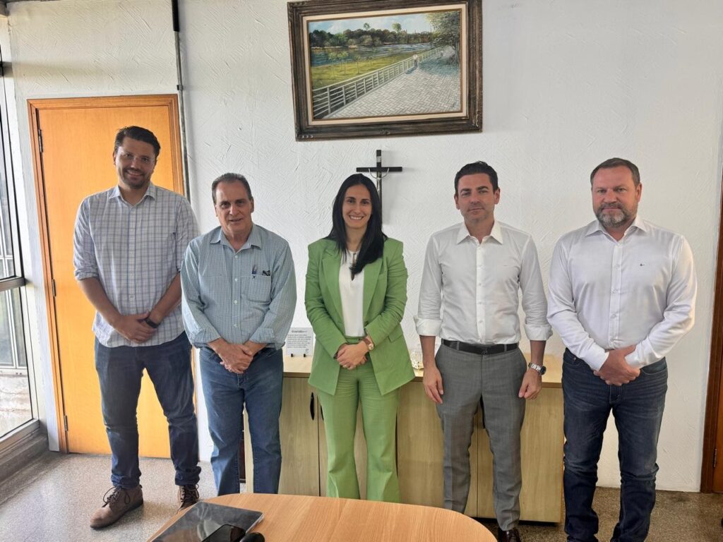 A imagem mostra um grupo de cinco pessoas, quatro homens e uma mulher, posando para uma foto em um ambiente interno. Eles estão em pé, alinhados frente a uma parede clara que tem um quadro pendurado, representando uma paisagem com árvores, um corpo d'água e uma cerca. Na parede também há um crucifixo. A iluminação parece ser natural, vinda do lado esquerdo, criando sombras suaves. As pessoas estão vestidas de forma casual e semi-formal: o homem à esquerda usa uma camisa xadrez e jeans, o segundo homem veste uma camisa azul clara e jeans, a mulher no centro está com um terno verde e blusa branca, o quarto homem usa uma camisa social branca e calças cinza, e o homem à direita veste uma camisa social branca e jeans escuros. Todos olham para a câmera com expressões neutras. No ambiente, além da parede com o quadro e o crucifixo, há uma porta de madeira à esquerda e um móvel em madeira ao centro, possivelmente um balcão ou armário. Na frente do grupo, há uma mesa de madeira com alguns objetos, incluindo um tablet. A foto tem boa qualidade, com foco nítido nas pessoas.
