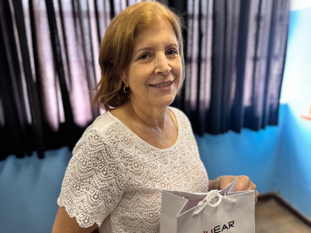 A imagem mostra uma mulher loira de meia-idade, sorrindo para a câmera. Ela está vestindo uma blusa branca com detalhes em renda nas mangas e no peito. Seus cabelos são curtos e levemente ondulados, com um tom acobreado. Ela usa brincos dourados em formato de argola. Ao fundo, há uma cortina escura com listras verticais, e uma parede azul com uma faixa colorida na parte inferior. A mulher está segurando uma sacola de presente branca com o logotipo "EAR" em letras vermelhas. Sua mão direita está apoiada na sacola, e ela usa uma pulseira verde no pulso. A iluminação parece ser natural, vindo de uma fonte de luz à sua direita.