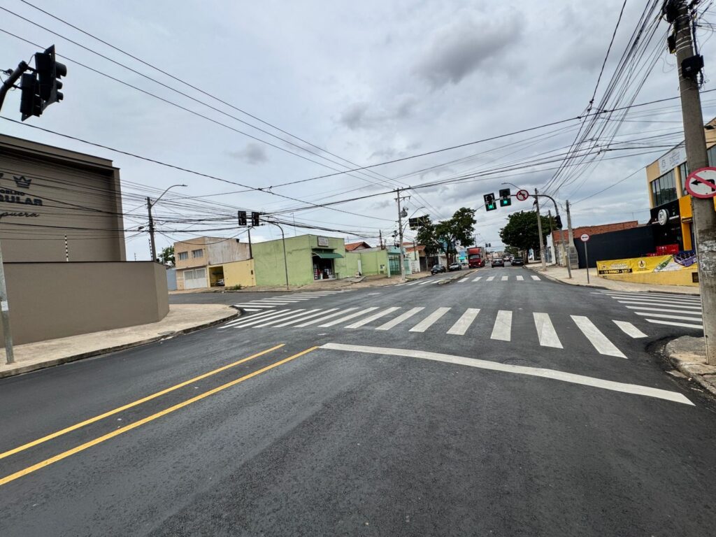 A imagem retrata um cruzamento de rua em um dia nublado. O asfalto da rua está recém-pavimentado, com marcações de faixa de pedestres e linhas amarelas duplas recém-pintadas. Vários edifícios de diferentes cores e tamanhos ladeiam a rua, incluindo um edifício cinza com o que parece ser um logotipo no canto superior esquerdo, um edifício amarelo e um edifício verde. Semáforos estão posicionados em vários pontos do cruzamento, e fios elétricos se estendem por todo o céu. Vários carros e um caminhão vermelho podem ser vistos na rua, com alguns carros parados em frente aos semáforos. Há também placas de trânsito visíveis, incluindo uma placa de "proibido virar à esquerda". O céu está nublado, com nuvens densas cobrindo o sol.